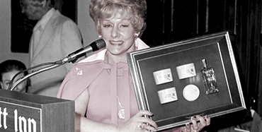 Mary Kay Ash smiles for a photo while standing at a podium and holding up a...